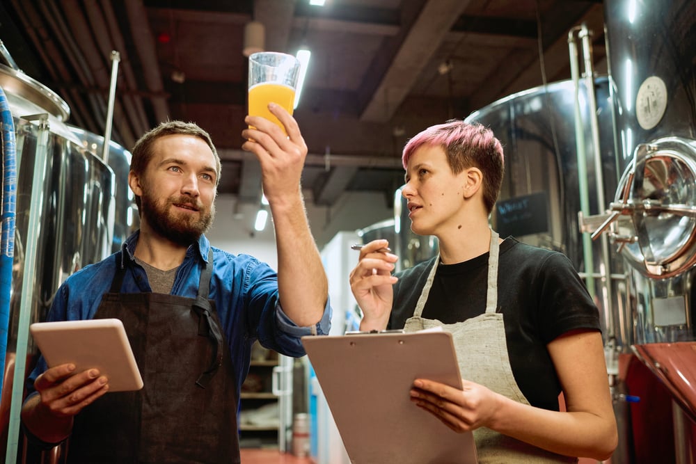 Two brewers in a brewery examine a hazy beer sample while holding clipboards beside stainless steel tanks.
